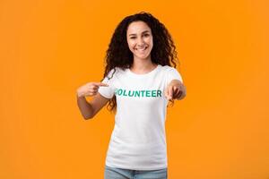 I'm a volunteer concept. Joyful female activist pointing at t-shirt uniform and at camera posing in studio over yellow background. Volunteering career, charity work photo