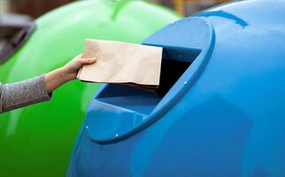 Waste Sorting. Unrecognizable Lady Throwing Paper Bag Into Blue Garbage Recycle Bin Outdoors, Woman Enjoying Separating Garbage, Caring About Environment, Cropped Image With Selective Focus photo