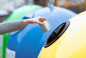 Garbage Sorting Concept. Woman hand throwing metal can into yellow recycle bin container outdoors, unrecognizable lady enjoying waste recycling system, cropped image with selective focus photo