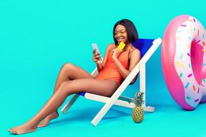 Carefree black woman holding credit card and smartphone, shopping online in lounge chair on blue background, panorama with free space. Sexy young lady buying goods in web store during summer vacation photo