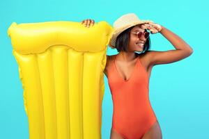Millennial black lady in swimsuit with inflatable lilo holding hand above her eyes, looking into distance on blue studio background. African American woman with pool float having great summer vacation photo
