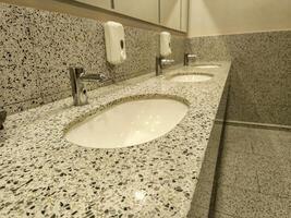 A bathroom with three sinks and a soap dispenser. The sinks are white and the counter is made of granite photo