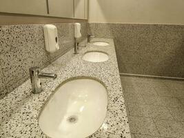 A bathroom with three sinks and a soap dispenser. The sinks are white and the walls are white photo