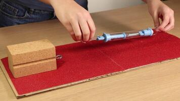 Hands use a spring scale to measure friction on wooden blocks over sandpaper. Close-up, well-lit, educational setup video