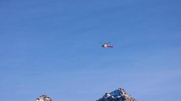 A helicopter flies across a clear blue sky above snow-capped mountains, capturing serene aerial movement in a natural landscape video