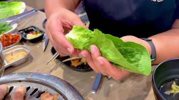 Hands assembling Korean BBQ wraps with lettuce and grilled meat in a vibrant Bangkok setting. Bright lighting enhances the culinary process video