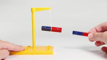 Hands manipulate magnets to show attraction and repulsion, using a suspended magnet setup. Bright lighting highlights the magnetic forces video