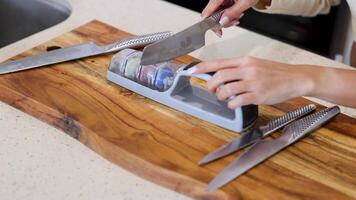 A person sharpens a knife using a manual sharpener on a wooden cutting board in a well-lit kitchen video