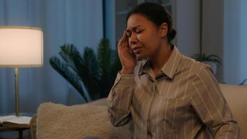 Sick African American woman use mobile phone gadget addict at night evening home ill unhealthy girl on couch feel headache migraine painful head cramp massaging temples suffer ache high blood pressure video