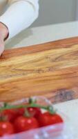 Hands wipe a wooden cutting board on a kitchen counter, with tomatoes nearby, in a well-lit setting video