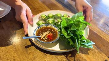 Hands arrange a Vietnamese dish with herbs and dipping sauce on a wooden table under natural light video