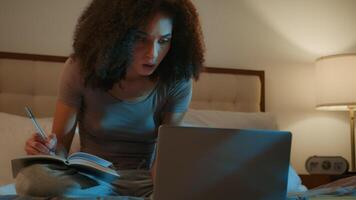 African American woman student high school learner pupil girl studying on bed use laptop watch lecture online prepare for exam night homework write notes in book learn subject e-learning at home video