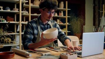 Caucasian man sculptor potter online laptop shopping in pottery workshop create handmade ceramics small business owner sale craft art male guy craftsman designer sell handcraft bowl crockery stoneware video
