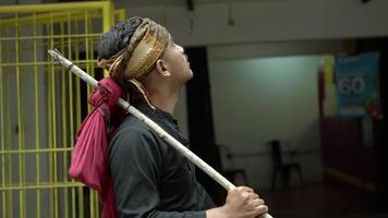 A man dressed in traditional attire, holding a stick and a red cloth draped over his shoulder. He looks contemplative, set against a blurred indoor background. video