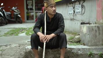 A man sitting on a stone step, wearing traditional black attire and a headscarf, holding a wooden stick. He appears contemplative, with a backdrop of urban elements and parked motorcycles. video