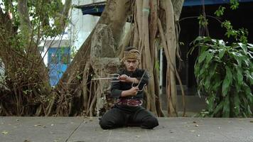 A martial artist in traditional attire performs a pose with weapons in a natural setting surrounded by trees and greenery. video