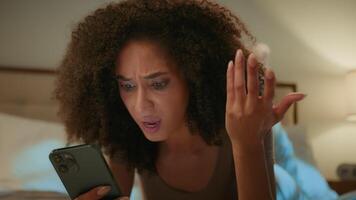 Shocked irritated girl African American young woman gen z user lying on bed with mobile phone at home evening night bedroom upset with bad news refusal loss connection failure smartphone problem fraud video