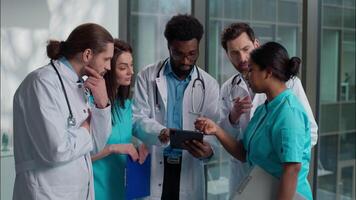 Group of five medical students diverse multicultural diagnostic team doctors men woman standing hospital lobby use tablet device nodding head agree prognosis healthcare rehabilitation discuss surgery video