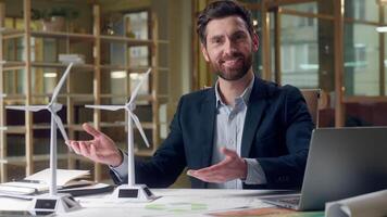 Successful smiling Caucasian business man presenting ecology project windmills green energy models laptop working generator power environment showing thumbs up raised fingers businessman male office video