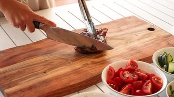 A person slices a beef steak on a wooden board beside fresh vegetables in bright, natural lighting video