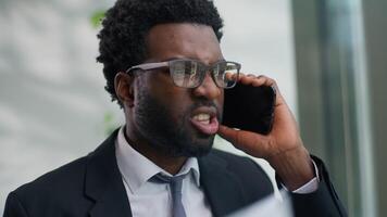 Angry mad African American business man yelling arguing talking mobile phone holding papers in office work documents mistake ethnic businessman male furious argument quarrel conflict smartphone call video