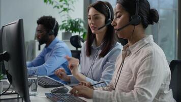 Cooperation Caucasian operator helping call center coworker Indian worker talking helpline women African men in headset typing computer writing notes callcenter support online trading sales at office video