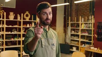 Happy glad smiling Caucasian man businessman male small business owner in apron seller potter pointing at camera index finger you showing thumb up gesture good great recommend indoor pottery workshop video