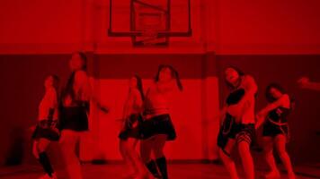 A group of six dancers performing in a gymnasium with a red lighting effect. They showcase various dance poses, wearing stylish outfits that reflect a modern dance aesthetic. video