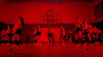 A dynamic dance performance in a gymnasium, featuring a group of dancers in coordinated outfits. The lighting is red, creating an energetic atmosphere. The dancers are in various poses video