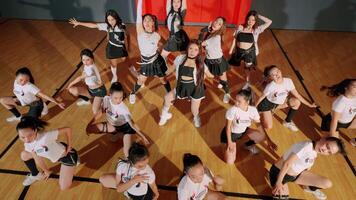 A group of young dancers performing a choreographed routine in a gymnasium. They are wearing matching outfits and showcasing various dance poses. The floor is wooden, and the background features video