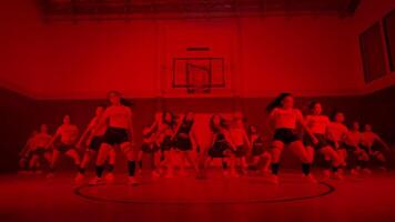 A group of dancers performing in a gymnasium with red lighting. The dancers are in various poses, showcasing their choreography. The basketball hoop is visible in the background. video