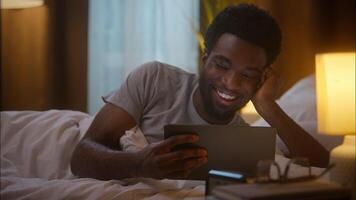 Joyful laughing African American ethnic man guy client male freelancer use digital tablet watching series funny social media content reading news touchscreen relaxing having fun at home bedroom video