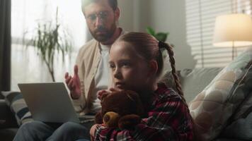 Caucasian family at home remote working dad using computer on sofa speaking to sad little girl child kid cuddling Teddy Bear conflict emotional stress childhood problem loneliness attention deficit video