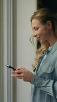 Happy woman client using mobile phone typing looking window waiting delivery at home Caucasian customer female businesswoman girl chatting smartphone standing wait deliverer ordering online app indoor video