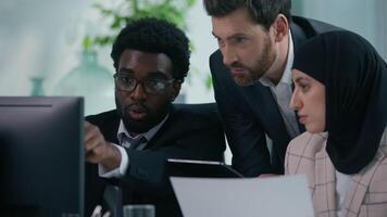 Three multiracial business colleagues in office with computer and paper document discuss startup project looking at screen multiethnic diversity manager coworkers talking working together cooperation video