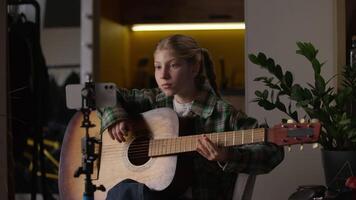 Little girl Caucasian child learning playing guitar watching online lesson tutorial at home focused kid schoolgirl blogger play musical instrument music song studying melody blog mobile phone tripod video