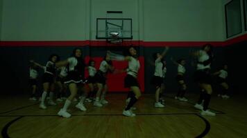 A group of dancers performing in a gymnasium with a basketball hoop. They are dressed in matching outfits, showcasing synchronized movements and energetic expressions. The lighting creates a dramatic video