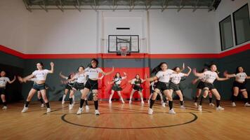 A group of dancers performing in a gymnasium, wearing matching outfits with a basketball hoop in the background. The choreography is energetic and synchronized, showcasing their skills. video