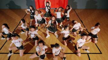 A vibrant dance group performing in a gymnasium, showcasing energetic choreography. The dancers wear matching outfits, featuring white tops and black skirts, with a dynamic arrangement. video