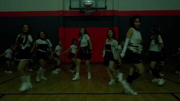 A dynamic dance performance in a gymnasium featuring a group of dancers in coordinated outfits. They are mid-jump, showcasing energy and synchronization, with a basketball hoop in the background. video