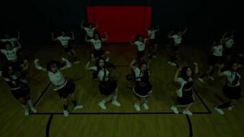 A group of dancers performing in a gymnasium, wearing matching outfits with skirts and sneakers. The scene is dimly lit, highlighting their synchronized movements and energetic poses. video