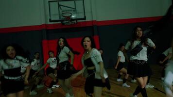 A dynamic dance scene in a gymnasium with dancers in motion. The lighting is low, creating a dramatic atmosphere. The focus is on a female dancer in the foreground, showcasing energy and movement. video