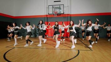 A group of dancers performing a choreographed routine in a gymnasium. They are dressed in matching outfits, showcasing energetic movements. The basketball hoop and court are visible in the background. video