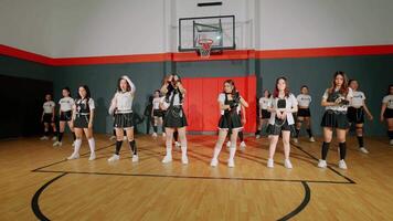 A group of young women performing a dance routine in a gymnasium. They are dressed in coordinated outfits, showcasing energy and teamwork. The background features a basketball hoop and court. video