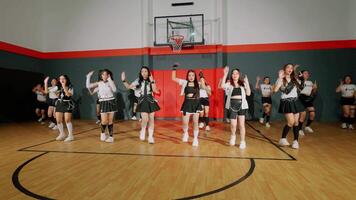 A group of young women performing a dance routine in a gymnasium. They are dressed in coordinated outfits, showcasing energetic movements. The basketball hoop is visible in the background. video