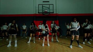 A group of dancers performing in a dimly lit basketball court. They wear coordinated outfits, showcasing a mix of sporty and stylish elements. The atmosphere is energetic and dynamic video