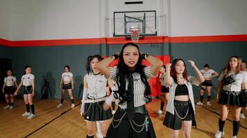 A dynamic dance performance in a gymnasium featuring a group of young women in stylish outfits. The central dancer stands confidently with hands on her head, surrounded by others video