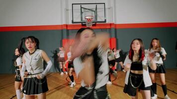 A dynamic dance performance in a gymnasium featuring multiple dancers in coordinated outfits. The scene captures energy and movement, with a basketball hoop in the background. video