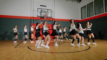 A group of dancers performing a choreographed routine in a gymnasium. They are wearing stylish outfits and showcasing energetic movements. The background features a basketball hoop and a wooden floor. video