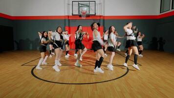 A group of dancers performing a choreographed routine in a basketball gym. They are wearing matching outfits and showcasing energetic movements. The gym features a basketball hoop and a wooden floor. video